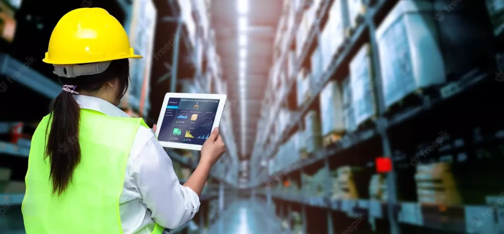system_oms Zdjęcie przedstawia kobietę w kasku i żółtej kamizelce na magazynie, która trzyma tablet przed sobą i analizuje dane na systemie WMS.